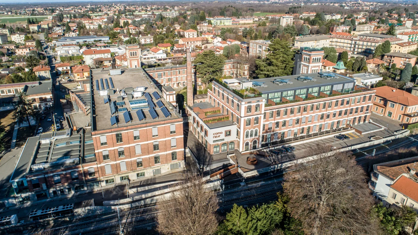 Vista aerea del campus ComoNExT a Lomazzo, con gli edifici in mattoni, la ciminiera e la città circostante.